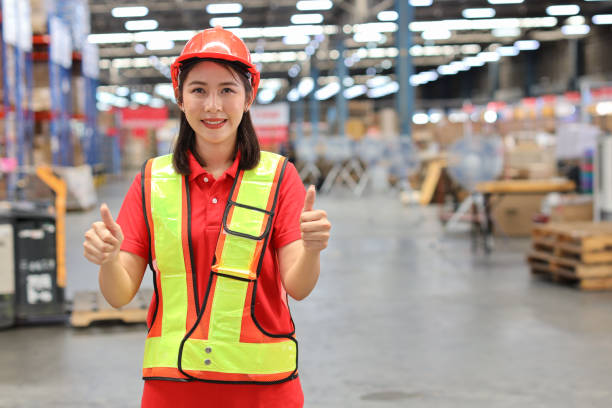istock Warehouse workers woman standing with hardhats while showing thumb up and looking at camera with production stock and retail warehouse logistics distribution center background 1494569385