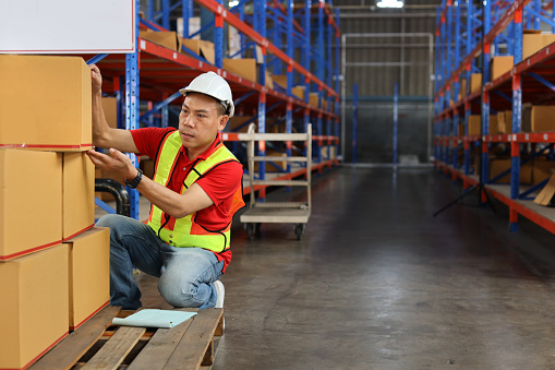 Warehouse worker man with saftyhat and reflective jacket checking list or control large box package for delivery to production stock and inventory in retail warehouse logistics, distribution center