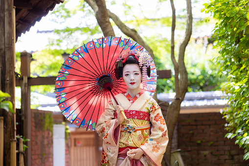Portrait of Japanese Maiko (Geisha in training) standing on narrow footpath in Gion, Kyoto, using traditional Japanese paper umbrella for sunshade
