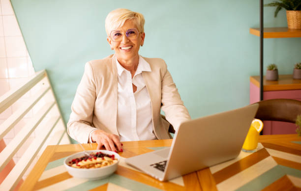 istock Senior woman using mobile phone while working on laptop and having healthy breakfast in the cafe 1494574921
