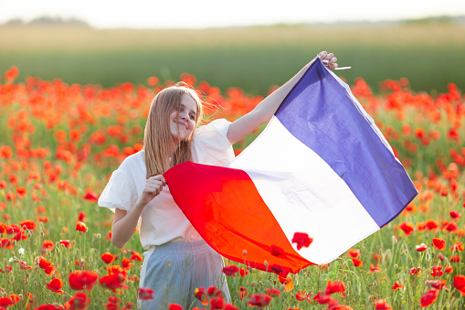 Beautiful girl holding waving flag of France in poppy field. National holidays concept: Armistice day, Bastille day. Travel and learn french language, beauty of France, french cosmetics and perfume.