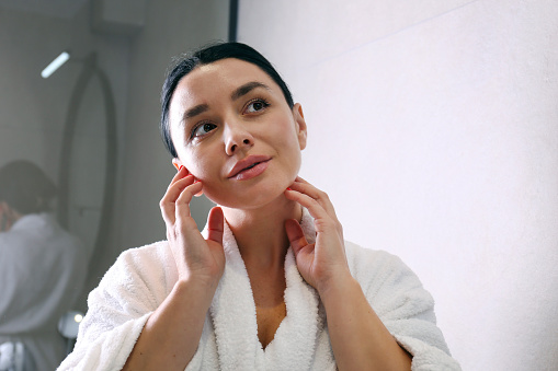 Young woman wearing a bathrobe looking at the round shaped mirror and applying anti-aging skincare to her face with massaging motions. Morning beauty routine concept. Close up, copy space, background.