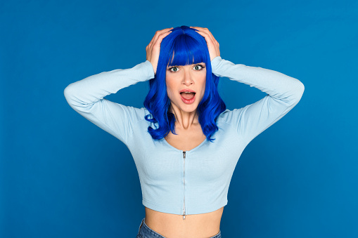Shocked young female model with dyed bright blue hair in casual clothes grabbing head and looking at camera with opened mouth in studio