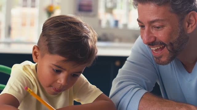 Close Up Of Mature Father At Home In Kitchen Helping Son With Homework