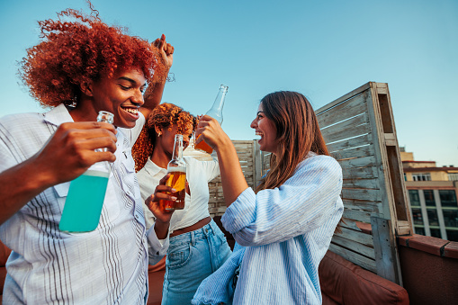 Three diverse young happy people dancing on the rooftop together while holding drinks and laughing