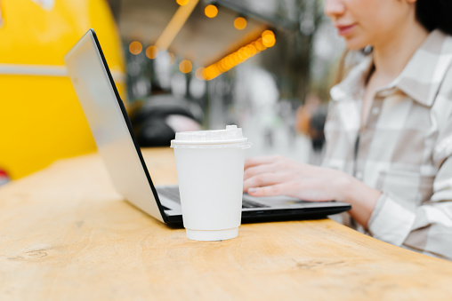 Close-up of woman working with laptop at street cafe table, freelancer holding typing keyboard during coffee break. Selective focus on a white disposable coffee cup.