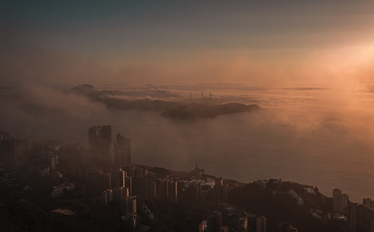 a birds eye view of a coastal city in silhouette and a nearby island across the sea during sunset with low clouds and sea fog rolling in and catching the golden sunlight under the clear sky