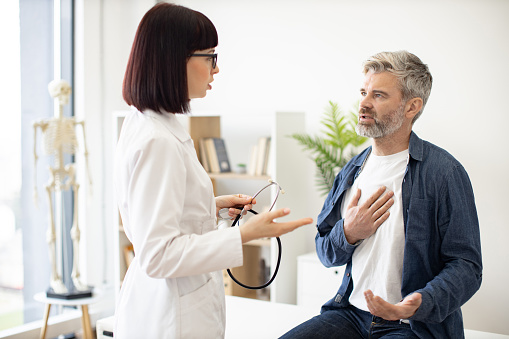 Worried gray-haired man sitting on exam couch with hand on chest while mindful woman in doctor's coat holding stethoscope. Male patient describing symptoms to medical specialist in consulting room.