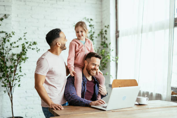 Mid adult gay couple with adopted daughter at home, working on laptop and having fun with girl stock photo