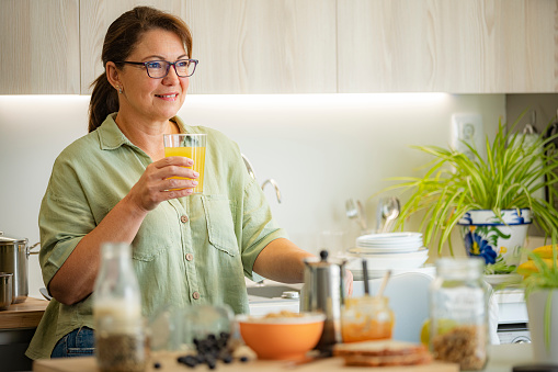 Smiling mature woman standing in kitchen drinking fresh organic fruit juice