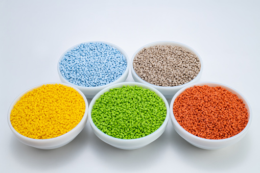 Creative composition of masterbatches, sky-blue, brown, yellow, green, and orange granules placed in five white bowls with white background, close-up shot.