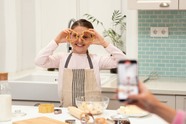 Caucasian young mom takes photo on smartphone of little daughter in apron, makes cookies, puts forms Caucasian young mom takes photo on smartphone of little daughter in apron, makes cookies, puts forms to eyes, has fun in kitchen interior. Cooking sweets, app for blog homemade food at home preschooler caucasian one person part of stock pictures, royalty-free photos & images