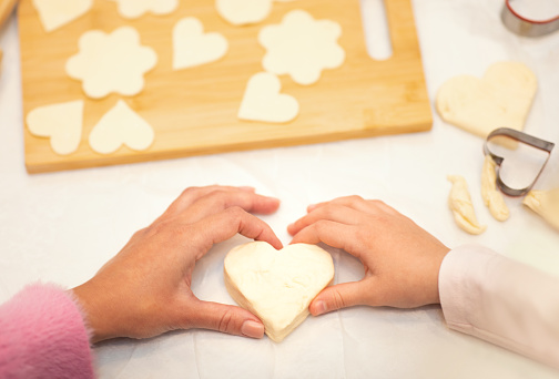 Hands of caucasian young mom and little daughter make cookies from dough in shape of heart on kitchen table with wooden board. Love homemade food, hobby, cooking sweets together at home, cropped