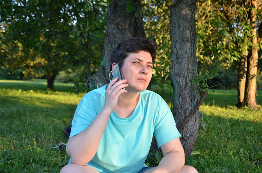 dark-haired woman with short hair in a blue T-shirt and shorts sits on the grass in the park, talking on a cell phone. woman during a telephone conversation.