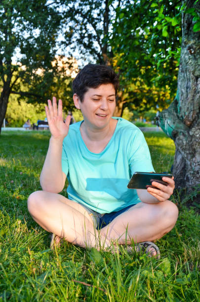 mujer sentada en el césped, sonriendo agitando la mano al interlocutor, hablando por videollamada en el teléfono inteligente - voip telecommunications equipment gratis freedom fotografías e imágenes de stock