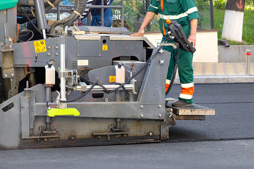 A road foreman stands on the footboard of an asphalt paver and controls the asphalt paving workflow with a remote control. Copy space.