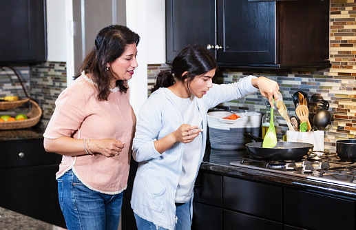 A teenage Indian girl cooking in the kitchen with her mother watching. The daughter is sauteing vegetables in a pan on a gas stove.