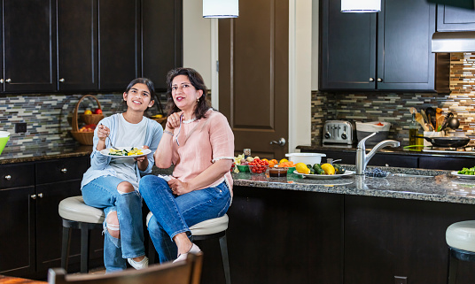 An Asian teenage girl and her mother sitting on kitchen stools, sharing a plate of sauteed vegetables. They are looking up, probably watching tv, smiling and pointing at it.