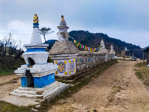 his captivating photograph showcases a stunning landscape adorned with a series of magnificent Stupas, or Manes, along the route to Sandakphu. Nestled against the backdrop of rolling hills, these religious monuments stand as remarkable testaments to the rich architectural heritage of Buddhism. The Stupas serve as sacred sites for worship and are also utilized as statues on graves, symbolizing the spiritual connection between life and death. Adding a vibrant contrast to the scene, a cascade of colorful prayer flags dances in the wind, infusing the image with a sense of tranquility and spirituality. As the beholder's gaze embraces the harmonious blend of natural beauty and cultural significance, this photograph invites contemplation and appreciation of the serene surroundings on the journey to Sandakphu.