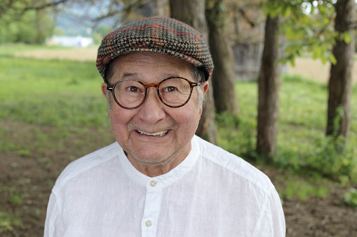 A senior Hispanic man in his seventies laughing out loud in nature. He wears an elegant white button down shirt with eyeglasses and hat.