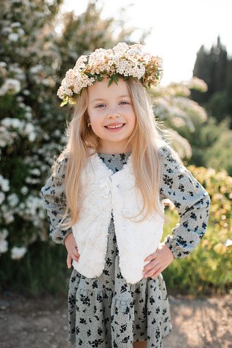 Smiling stylish child girl 4-5 year old wear floral wreath hairstyle and dress standing over flower at background outdoor. Look at camera. Summer season.