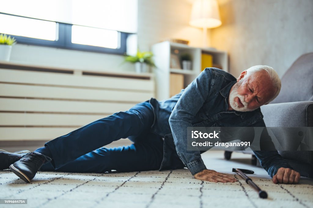 Old man suffering with pain and struggling to get up after falling down at home A Senior Man who is Using a Walking Stick is Lying on the Wooden Floor in his Apartment After he Fell in Serious Accident. Senior man lying on the floor of his room in an assisted living home Falling Stock Photo