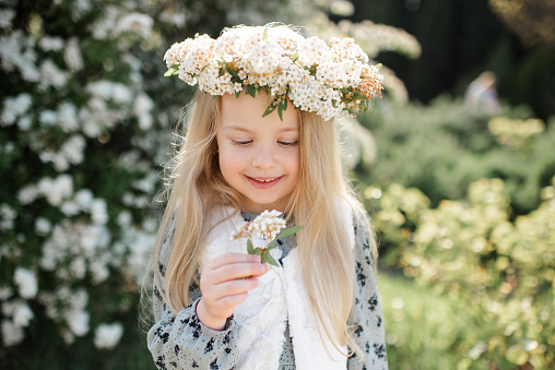 Smiling stylish child girl 4-5 year old wear floral wreath hairstyle and dress standing over flower at background outdoor. Summer season.