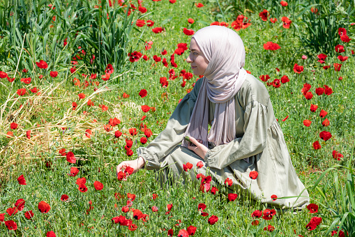 religious woman in hijab in long dress among poppy flowers. In the spring season, red poppy flowers rose from the green grass. woman watching the beauty of nature. Shot with a full-frame camera on a sunny day.