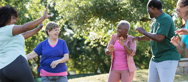 A multiracial group of five adults having fun dancing and exercising at the park on a sunny day. The focus is on the two senior women in the middle, smiling and laughing, looking toward each other. They are in their 70s.