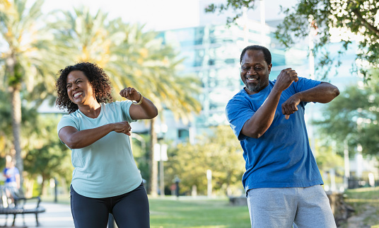 A mature Hispanic couple having fun dancing and exercising outdoors in a city park. The African-American man and Hispanic woman are standing side by side, looking at each other, smiling and laughing.