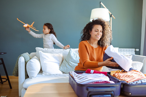 Latin woman smiling packing clothes in suitcase. Her little daughter is playing with airplane toy in background