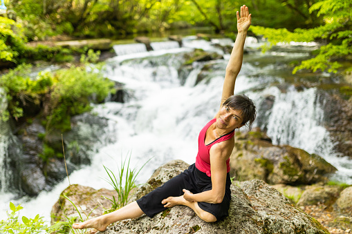 A mid adult woman enjoying yoga stretching relaxation in front of a waterfall.