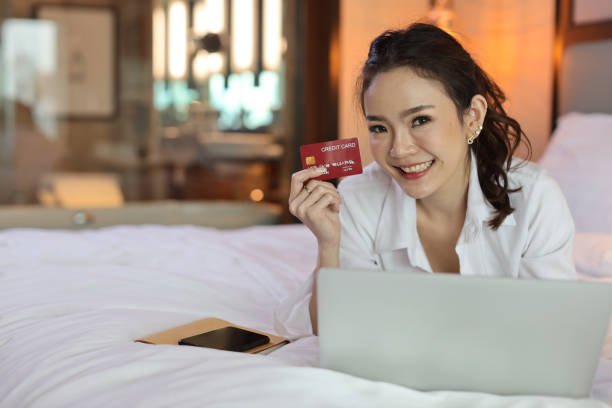 istock Happy asian woman in casual lying on bed and holding credit card while using computer in hotel for online shopping payment, beauty businesswoman with bedroom background. Lifestyle technology concept 1494669556