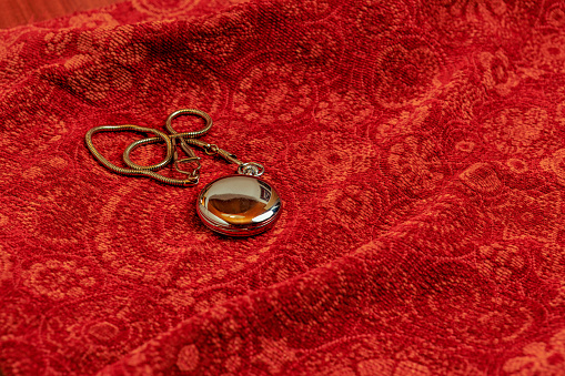 A shiny pocket watch has been placed on a rich red upholstery fabric. It is made of Stainless steel and is buffed to shine at its best. It has a matching stainless steel chain. Indoor studio shot; copy space. No people.