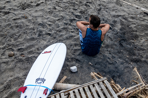 Canggu, Bali, Indonesia, May 10, 2023 A man on the dark sand Canggu beach sits next to a surfboard.