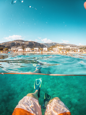Underwater shot with view of the coastline in the background. Adult wearing scuba diving equipment enjoys a sunny day of exploration.