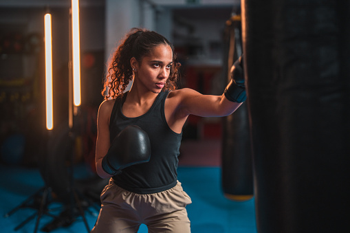 Three-quarter length shot of a young multiracial female boxer punching a bag. She is indoors in a gym, wearing sports clothing and boxing gloves.