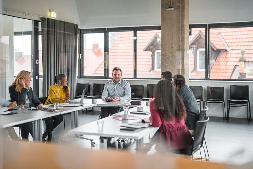 Through window shot of team, including a young man on a wheelchair, seated around a white table in a bright office with large windows. They are collaborating and working together.