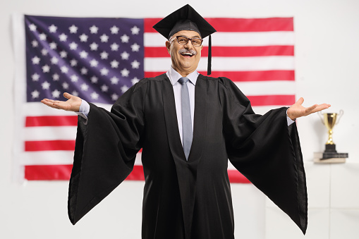Cheerful mature man wearing a graduation gown in front of the USA flag