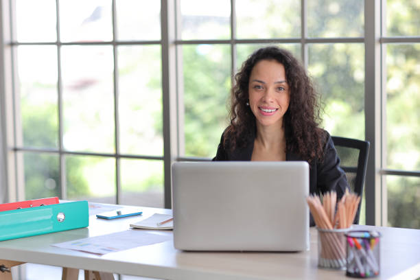 istock Attractive professional latin female employee worker sitting, using laptop computer with smart mobile phone at home workplace. Businesswoman working on paperwork while looking at camera and smile 1494683725