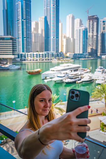 A young woman takes a selfie with her mobile phone on the Dubai Marina Promenade, holding a glass of champagne.