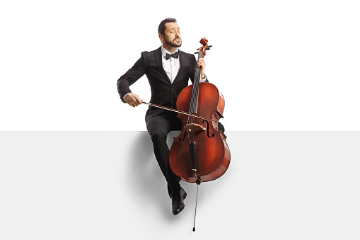 Male musician in a black suit and bow-tie sitting on a blank panel with a cello isolated on white background