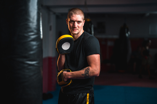Waist up shot of an athletic  mid adult Caucasian male boxing trainer using boxing equipment. He is indoors in a boxing gym, looking at the camera.