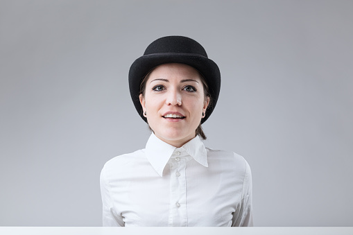Young woman with bowler hat, semi-open lips, speaking to you. Half-length, seated, neutral background. Undeniable beauty