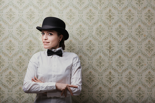 Against a retro yellow-green backdrop, a young woman dons a white shirt, bow tie, and bowler hat. Her arms are crossed, she sports a short haircut, and gives an expectant, inquiring smile