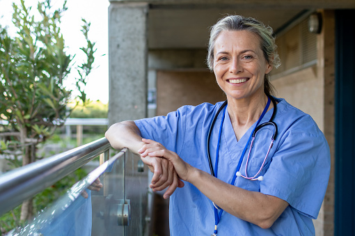 A caucasian female GP, standing in front of a hospital. Looking directly at the camera and smiling. Wearing blue medical scrubs with a stethoscope around her neck. Leaning on a balcony railing.