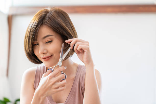 retrato de belleza mujer asiática tiene tijeras cortando el cabello a mano durante el confinamiento por coronavirus. la chica tiene un nuevo corte de pelo peinado, corte de pelo de bricolaje durante covid quédate en casa, nuevo concepto de peluquería de -   brunette  fotografías e imágenes de stock