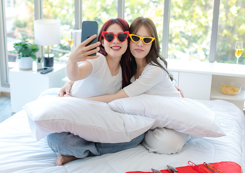 Asian young cheerful female LGBTQ lesbian lover couple partner smiling trying funny sunglasses together smiling sitting on bed taking selfie photo with smartphone with rainbow equality freedom flag.