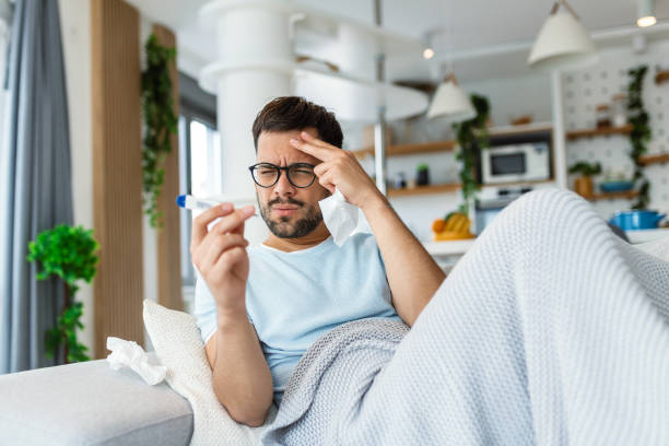 istock Man feeling sick lying in the bed and looking the thermometer. Sick man lying on sofa checking his temperature under a blanket at home in the living room 1494730831