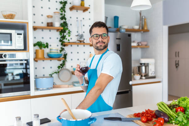 istock Home cook. Single man in apron looking at the window, drinking wine from a glass while cooking in the kitchen, cooking at home 1494730936